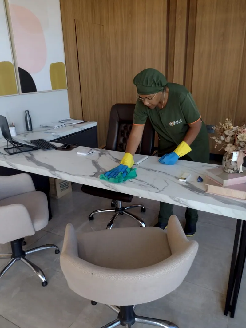 Funcionária da Solve, de uniforme verde e luvas, limpando uma mesa de mármore num escritório